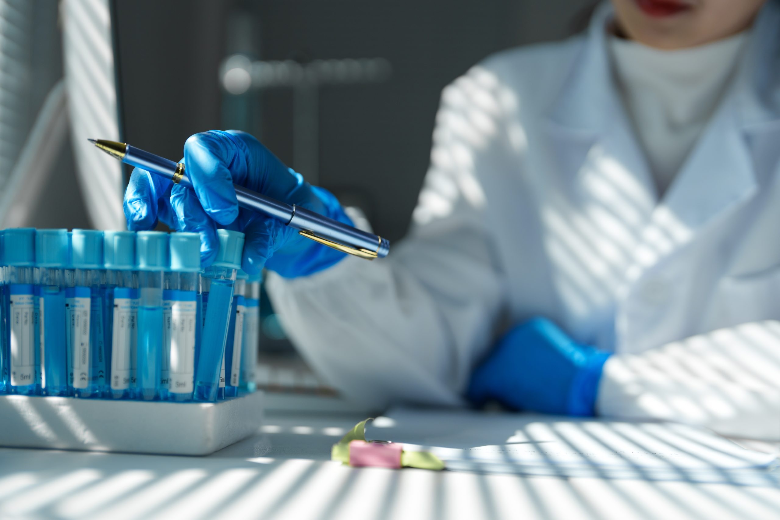 Medical researcher wearing gloves pointing with a pen at test tubes containing blue liquid, working on innovative research in a modern laboratory, backlit by window blinds